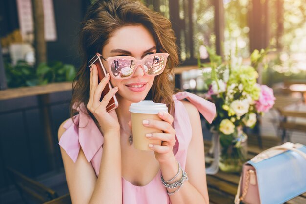 Retrato de uma jovem mulher atraente, sentado no café, roupa de moda de verão, vestido de algodão rosa, óculos de sol, sorrindo, bebendo café, acessórios elegantes, roupas da moda, falando no telefone