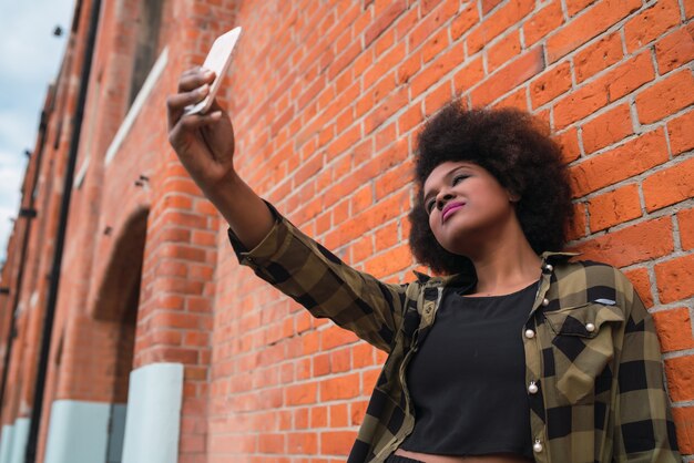 Retrato de uma jovem mulher bonita afro-americana do latim tomando uma selfie com seu telefone móvel ao ar livre na rua.