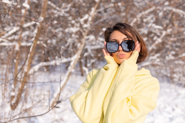 Foto grátis retrato de uma jovem mulher bonita em uma floresta de inverno com neve em um dia ensolarado, vestida com uma grande camisola amarela, com óculos de sol, aproveitando o sol e a neve