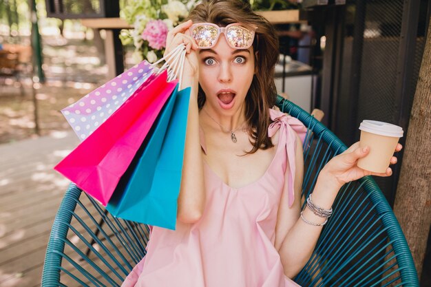 Retrato de uma jovem mulher bonita feliz sorrindo com expressão de rosto surpreso, sentado no café com sacolas de compras, bebendo café, roupa de moda verão, vestido de algodão rosa, vestuário da moda