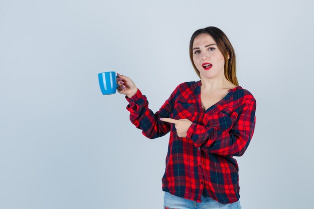 Retrato de uma jovem mulher bonita segurando a xícara enquanto aponta para a esquerda em uma camisa casual, jeans e uma bela vista frontal