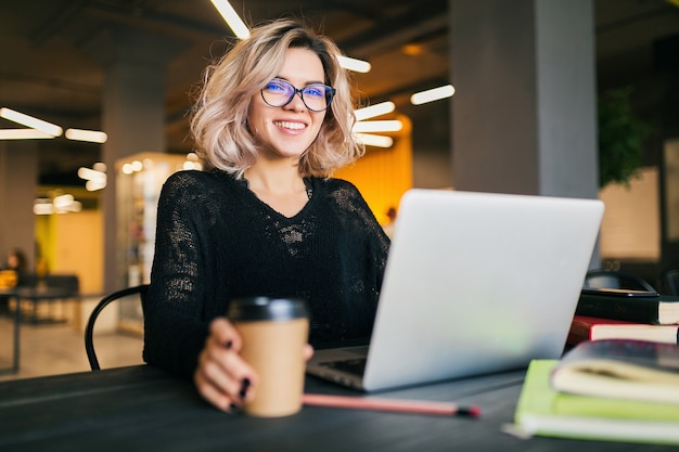 Retrato de uma jovem mulher bonita sentada à mesa com uma camisa preta, trabalhando no laptop em um escritório colaborativo