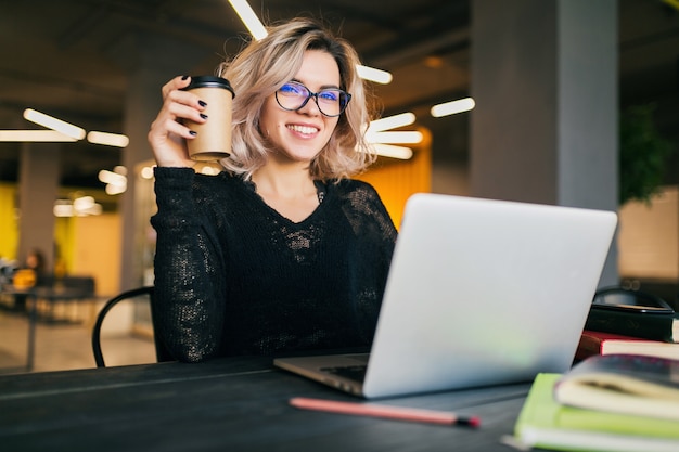 Retrato de uma jovem mulher bonita sentada à mesa na camisa preta, trabalhando no laptop no escritório colaborador, usando óculos, sorrindo, feliz, positivo, bebendo café em copo de papel