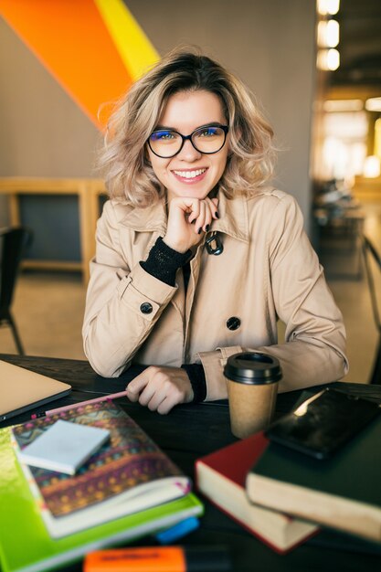 Retrato de uma jovem mulher bonita sentada à mesa no casaco trabalhando no laptop no escritório colaborador, usando óculos, sorrindo, feliz, positivo, local de trabalho