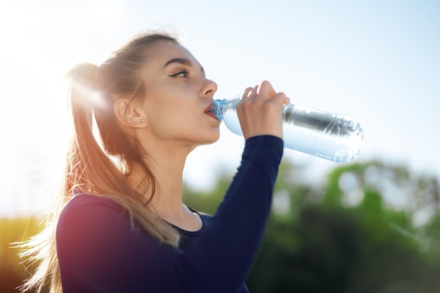 Retrato de uma jovem mulher bonita vestindo roupas esportivas azuis bebendo água no parque