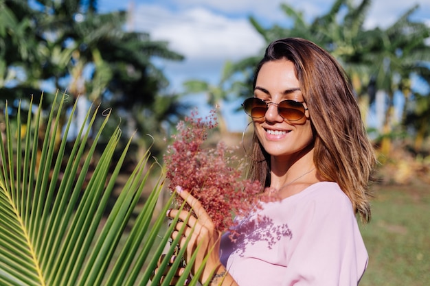 Retrato de uma jovem mulher bronzeada caucasiana em um vestido rosa romântico, pulseira de prata com brincos redondos e óculos escuros com flores silvestres