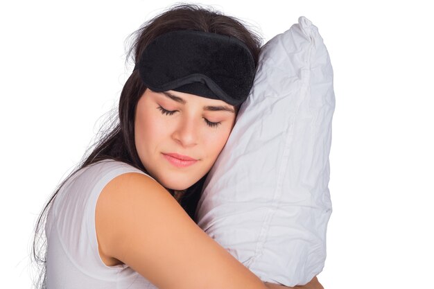 Retrato de uma jovem mulher cansada com máscara de dormir, descansando e segurando uma almofada no estúdio.