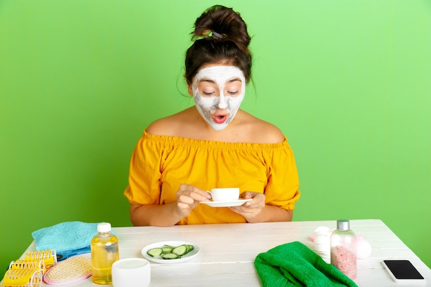 Retrato de uma jovem mulher caucasiana na rotina de cuidados de dia, pele e cabelo da beleza. Modelo feminino, bebendo café, chá enquanto aplica a máscara facial. Atônito. Conceito de autocuidado, beleza natural e cosméticos.