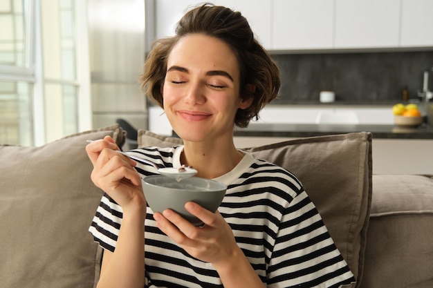 Foto grátis retrato de uma jovem mulher comendo uma tigela de cereais com leite sentada no sofá e tendo seu