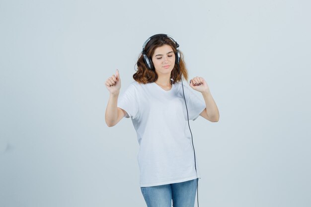 Retrato de uma jovem mulher curtindo música com fones de ouvido em uma camiseta branca, jeans e olhando para a frente alegre