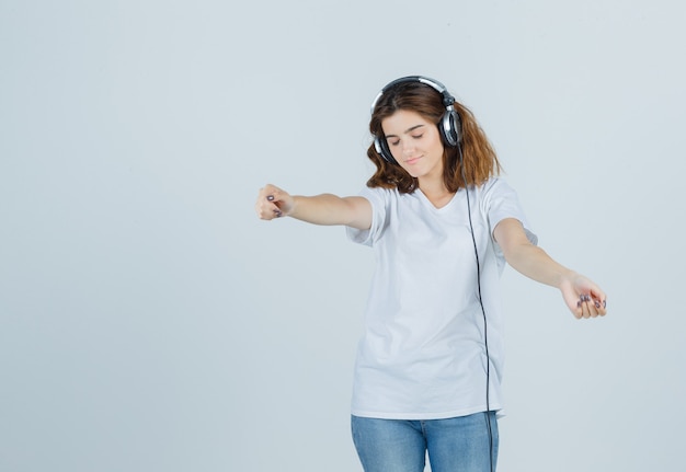 Retrato de uma jovem mulher curtindo música com fones de ouvido em uma camiseta branca, jeans e uma vista frontal divertida