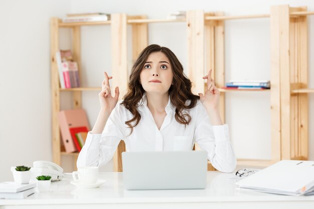 Retrato de uma jovem mulher de negócios caucasiana feliz sentada na mesa de escritório e segurando os dedos cruzados isolados sobre fundo branco