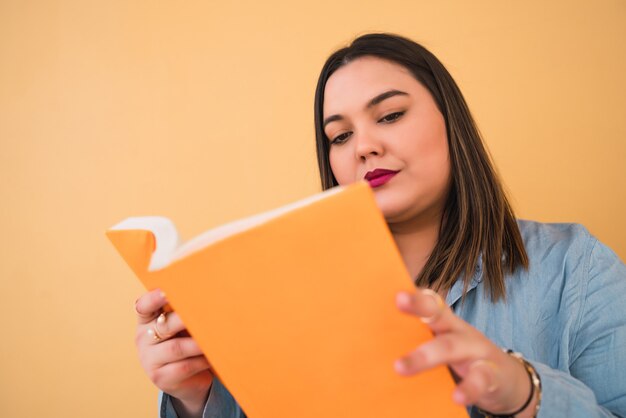 Retrato de uma jovem mulher de tamanho, curtindo o tempo livre e lendo um livro em pé contra a parede amarela.