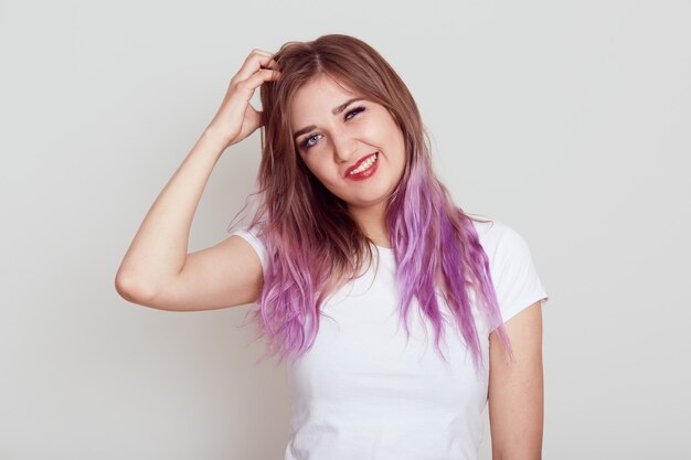 Retrato de uma jovem mulher em uma camiseta mais branca de estilo casual, coçando o cabelo de caspa e irritação, sofrendo de piolhos, rosto carrancudo, isolado sobre fundo cinza.