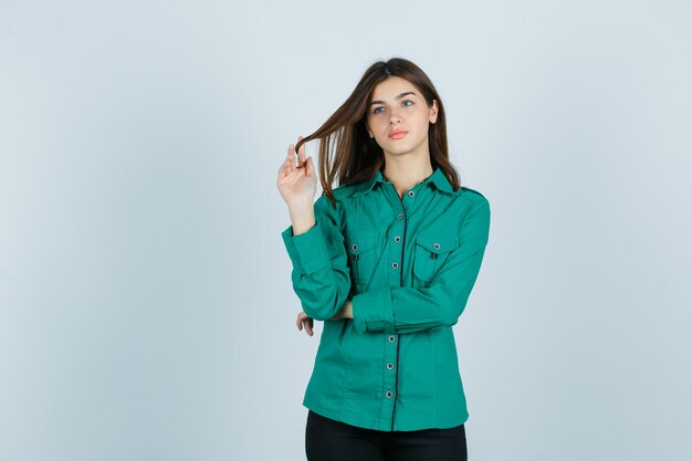 Retrato de uma jovem mulher enrolando o cabelo castanho em volta dos dedos em uma camisa verde e olhando a vista frontal atenciosa