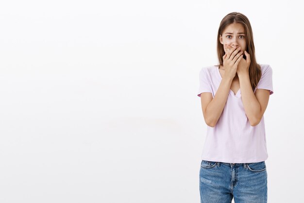 Retrato de uma jovem mulher europeia chateada e apavorada cobrindo a boca com as duas mãos franzindo a testa e gritando de medo sobre uma parede branca