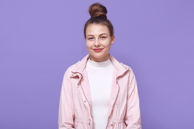 Retrato de uma jovem mulher feliz isolada parede lilás, vestindo jaqueta rosa pálida e camisa branca, tem coque de cabelo, senhora com aparência agradável.