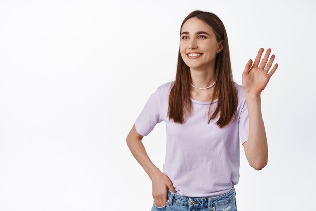 Retrato de uma jovem mulher moderna acenando olá, olhando de lado para as pessoas e sorrindo, cumprimentando, vendo um amigo e acenando com um gesto de olá, de pé no branco