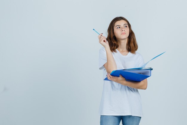 Retrato de uma jovem mulher segurando pastas e caneta em uma camiseta branca, jeans e olhando pensativa para a frente