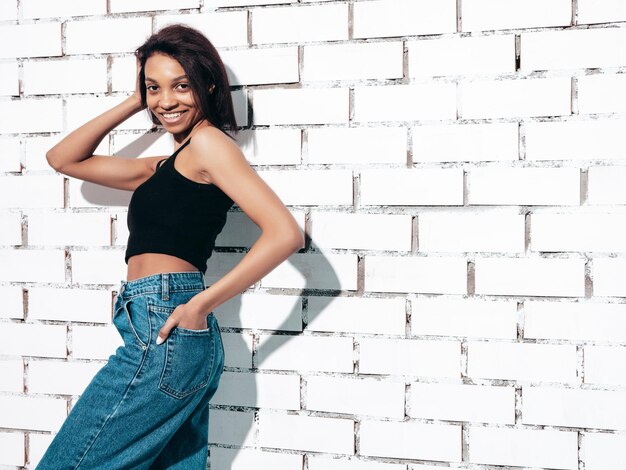 Retrato de uma jovem negra linda modelo sorridente vestido com jeans de verão e roupas pretas de cima Mulher sexy despreocupada posando perto da parede de tijolos brancos no estúdio Bronzeado e alegre