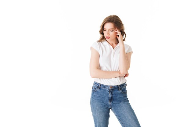Retrato de uma jovem pensativa de pé em camiseta e jeans e fechando cuidadosamente os olhos em fundo branco isolado