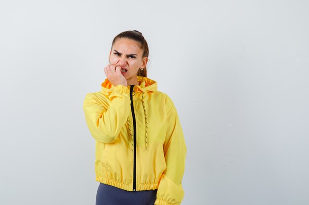 Retrato de uma jovem roendo unhas em uma jaqueta amarela e olhando a vista frontal nervosa
