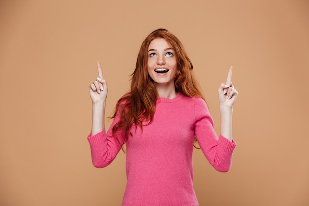 Retrato de uma jovem ruiva alegre apontando para cima com os dedos