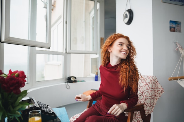 Retrato de uma jovem ruiva feliz sentado em uma cadeira à mesa com a máquina de escrever em casa