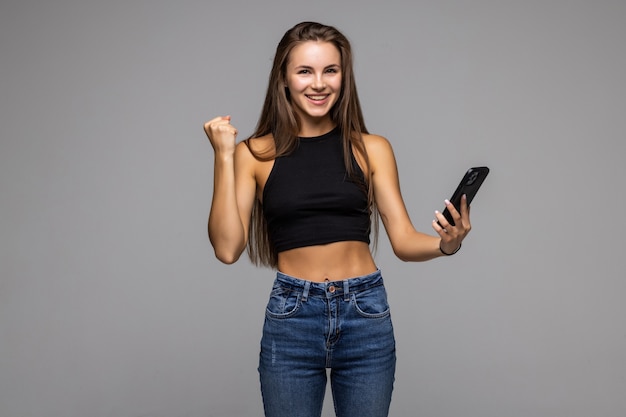 Retrato de uma jovem satisfeita segurando um telefone celular e comemorando isolado em um fundo cinza