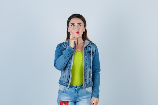 Retrato de uma jovem segurando o dedo na bochecha enquanto olha para cima com uma roupa jeans e olhando pensativa para a frente