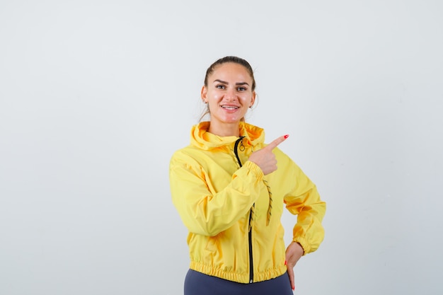 Retrato de uma jovem senhora apontando para o canto superior direito com um agasalho esportivo e olhando de frente com alegria