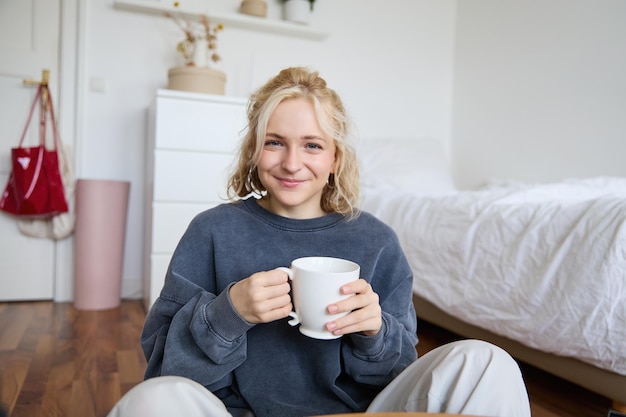 Foto grátis retrato de uma jovem sentada no chão do quarto bebendo chá segurando uma caneca branca e sorrindo