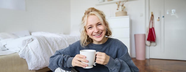 Foto grátis retrato de uma jovem sentada no chão do quarto bebendo chá segurando uma caneca branca e sorrindo