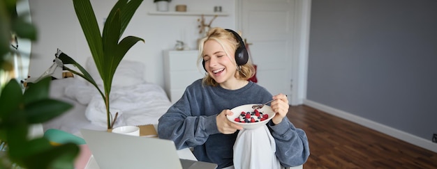 Retrato de uma jovem sorridente a ver um programa de televisão com fones de ouvido a tomar o pequeno-almoço e a olhar para