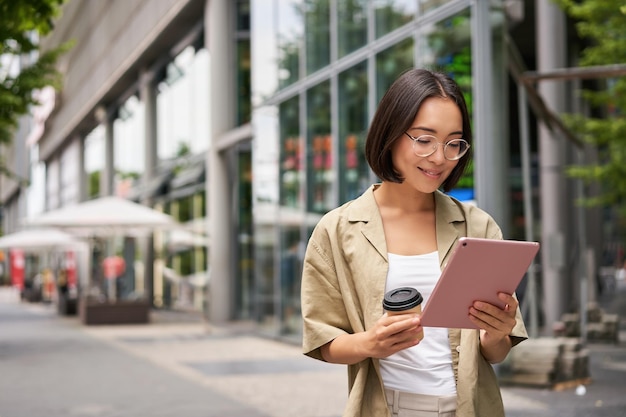 Retrato de uma jovem sorridente andando na cidade com um tablet bebendo café para viagem descendo o