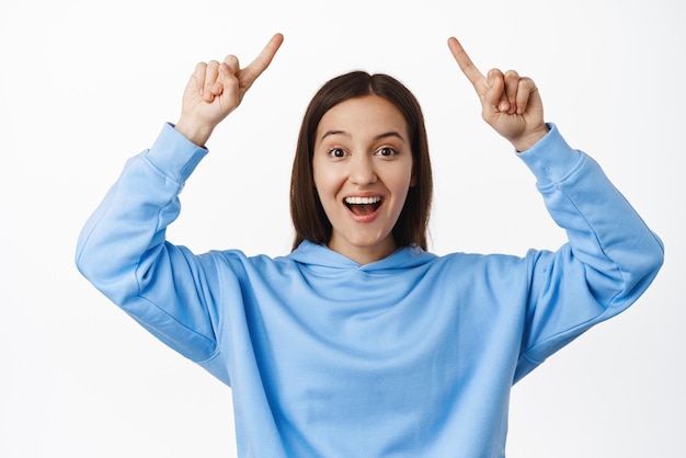 Retrato de uma jovem sorridente, apontando os dedos para cima no logotipo da venda, mostrando o anúncio do programa de publicidade de texto de promoção em pé no capuz contra fundo branco