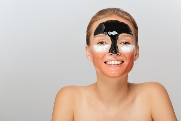 Retrato de uma jovem sorridente com diferentes máscaras cosméticas no rosto olhando alegremente na câmera sobre fundo cinza isolado