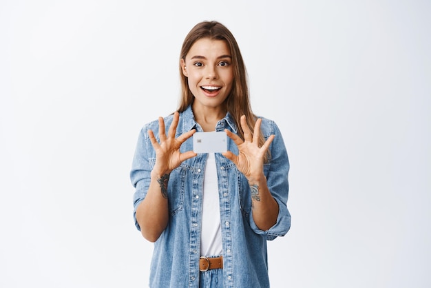 Foto grátis retrato de uma jovem sorridente e surpresa segurando o cartão de crédito plástico nas mãos sobre o peito anunciando novo recurso bancário recomendando fundo branco do banco