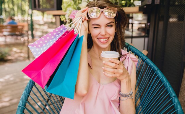 Retrato de uma jovem sorridente feliz mulher bonita com expressão de rosto animado, sentado no café com sacolas de compras bebendo café, roupa da moda para o verão, estilo hippie, vestido rosa de algodão, roupas da moda