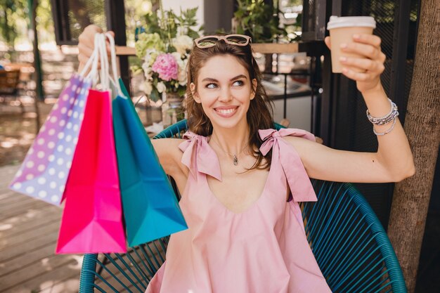 Retrato de uma jovem sorridente feliz mulher bonita com expressão de rosto animado, sentado no café com sacolas de compras bebendo café, roupa da moda para o verão, estilo hippie, vestido rosa de algodão, roupas da moda