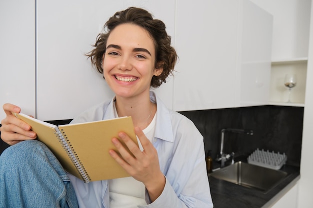 Retrato de uma jovem sorridente lendo um diário, desfrutando do conforto em casa, segurando um caderno e parecendo feliz.