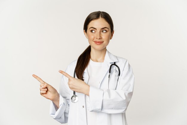 Retrato de uma jovem sorridente, médica, profissional de saúde, apontando os dedos para a esquerda, mostrando c...