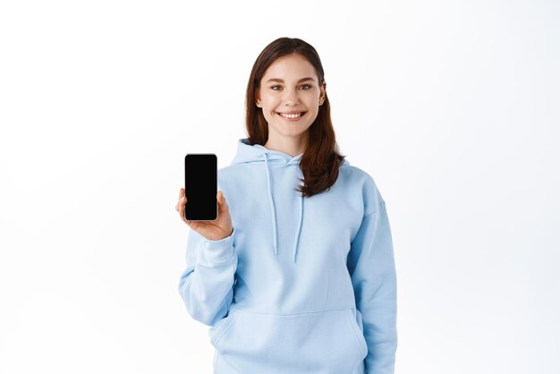 Retrato de uma jovem sorridente mostrando o celular de tela em branco isolado sobre fundo branco