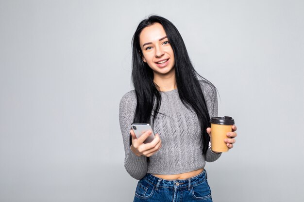 Retrato de uma jovem sorridente na mensagem de mensagem de camisa no telefone celular e segurando a xícara de café para ir isolado na parede cinza