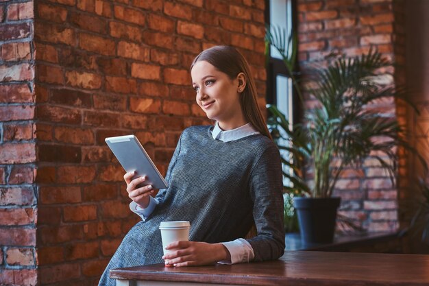 Retrato de uma jovem sorridente, vestida com um elegante vestido cinza detém uma xícara de café para viagem e tablet enquanto se inclina sobre a mesa em uma sala com interior loft, olhando para longe.