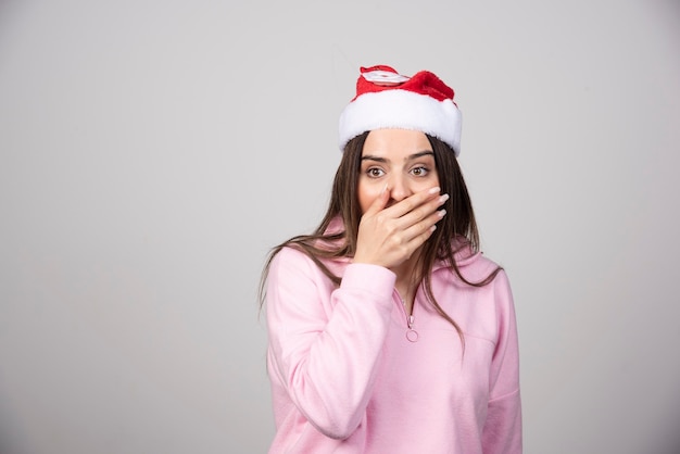 Retrato de uma jovem surpresa com chapéu de Papai Noel, cobrindo a boca com a mão.