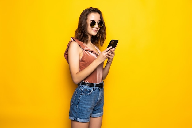 Retrato de uma jovem surpresa com chapéu de verão e óculos de sol, usando telefone celular isolado sobre a parede amarela