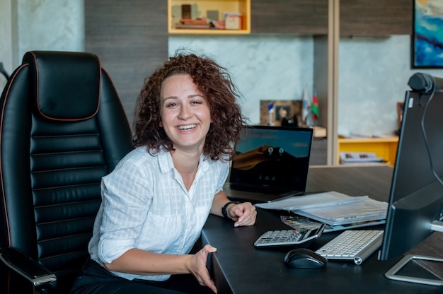 Foto grátis retrato de uma jovem trabalhadora de escritório sentada na mesa de escritório usando um laptop, olhando para a câmera, sorrindo alegremente trabalhando em um escritório luminoso