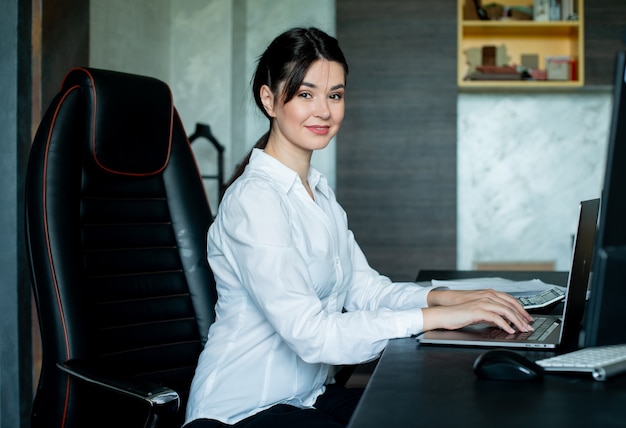 Retrato de uma jovem trabalhadora de escritório sentada na mesa do escritório, usando um laptop, olhando para a câmera, sorrindo alegremente, trabalhando no escritório