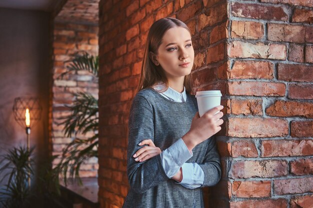 Retrato de uma jovem vestida com um elegante vestido cinza encostado em uma parede de tijolos segura uma xícara de café para viagem olhando para longe.